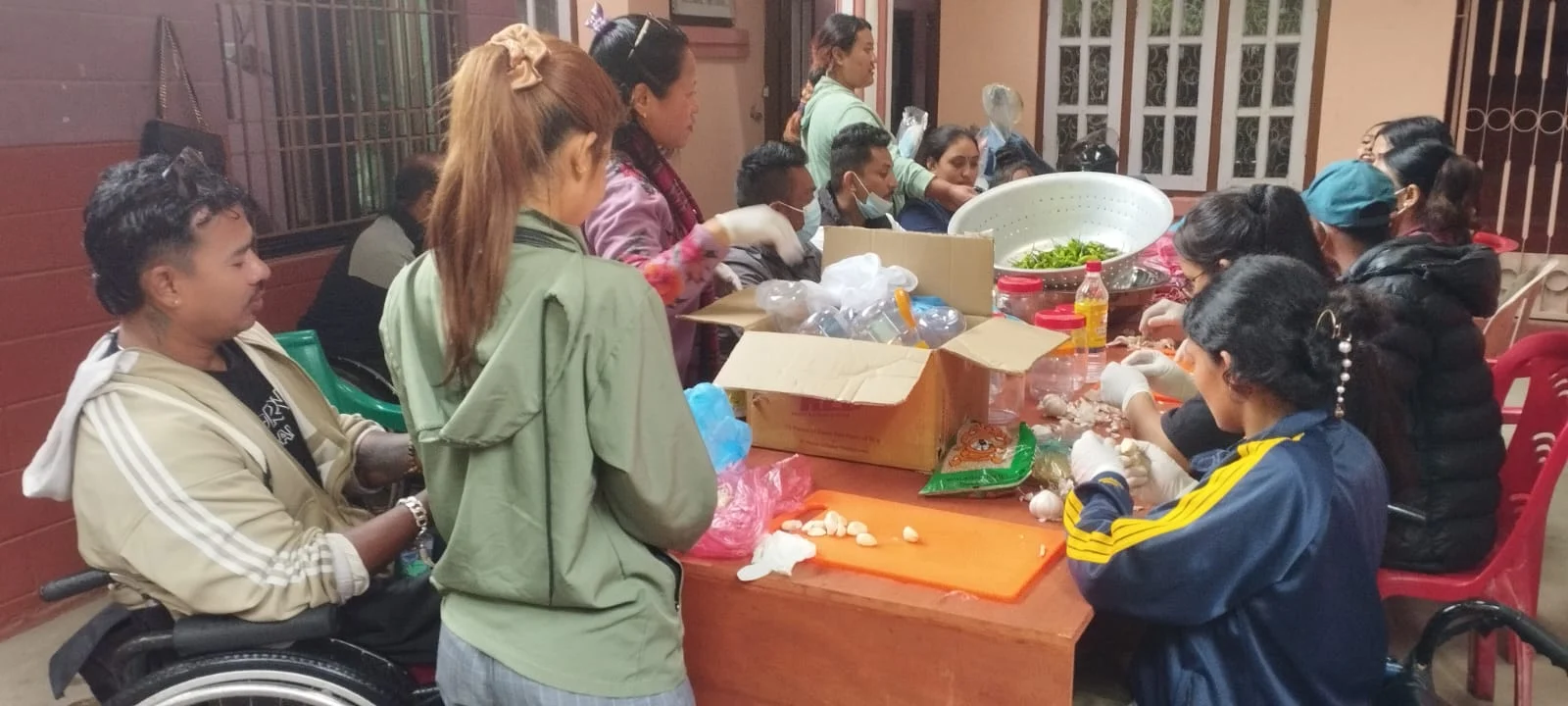 Persons with disabilities are participating in a pickle-making training session at the SCINN office in Makkalbari, Kathmandu. Seated around a table, they are peeling garlic and preparing ingredients like green chilies and oil, under the supervision of trainers.
